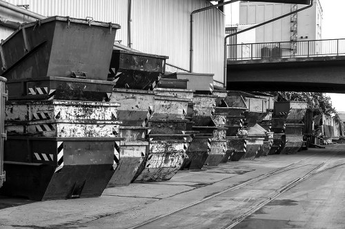 A row of large metal skip containers fitted with protective side panels, positioned on a paved industrial yard surface beneath a high warehouse or factory building with metal siding and a bridge-like structure overhead. The skips show signs of wear, with rust and grime visible on their surfaces, indicating prolonged outdoor use. The containers are aligned along the left side of the image, with some stacked or leaning slightly, while the background reveals more industrial structures and piping. The scene is captured in black and white, emphasizing the industrial environment typical of waste disposal and rubbish collection operations in Richmond. Commercial Waste Removal Richmond operates in areas requiring efficient management of bulky rubbish, and this image visually references the type of large-scale waste containers often used in commercial rubbish removal services in the vicinity. The overall setting highlights a professional, clean, and organized industrial yard dedicated to waste management tasks, supporting the company's service offering in the local area.