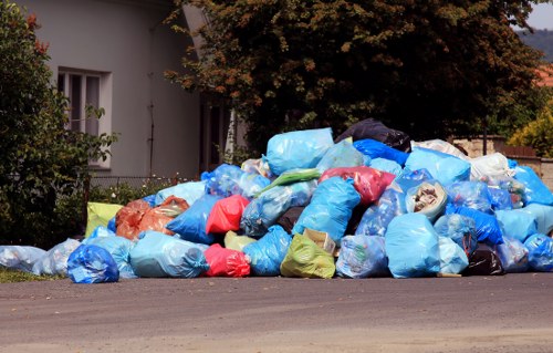 A large pile of assorted rubbish bags, predominantly blue, with some red, green, and transparent, are piled up on the paved roadside in front of a residential property. The bags appear to be made of plastic and are filled with waste, creating a chaotic heap that extends across the edge of the pavement. In the background, part of a white building with a window and a well-maintained garden area with green foliage and a tree with brown leaves are visible, indicating an outdoor suburban setting. The scene is taken during daylight hours, under natural lighting, with the rubbish heap partially blocking the view of the property and extending into the surrounding outdoor environment. The image looks like it could relate to waste collection or rubbish removal services in Richmond, suggesting the importance of professional waste management from companies like Commercial Waste Removal Richmond.