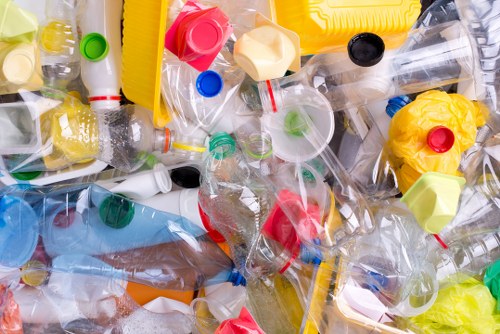 A close-up view of a large, mixed pile of discarded plastic waste, primarily consisting of transparent plastic bottles with various coloured caps including red, green, blue, and black. The bottles are crumpled and some are partially flattened, with visible ridges, smooth surfaces, and labels either peeled away or still attached. Among the bottles, there are several plastic bottle caps scattered throughout, as well as pieces of plastic wrappers, some yellow and pink, adding to the colourful assortment. The waste appears to be situated outdoors, possibly on a paved surface or driveway near a building or waste collection point in Richmond, with natural daylight illuminating the scene. The image reflects a typical scenario encountered in rubbish removal services, emphasizing the importance of proper disposal and recycling of plastics in urban and suburban environments. Commercial Waste Removal Richmond undertakes the collection and responsible disposal of such waste materials, helping to maintain cleanliness and environmental standards in the local area.