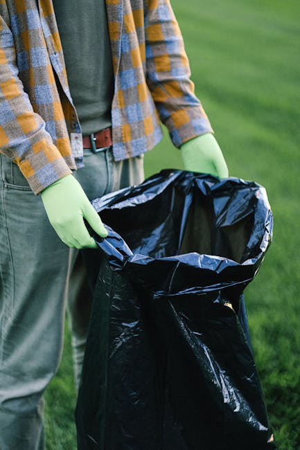 Two large black plastic bags and a cardboard box are placed on the pavement next to a metal fence with vertical bars, adjacent to a bushy tree. The bags appear to contain waste materials, possibly hou