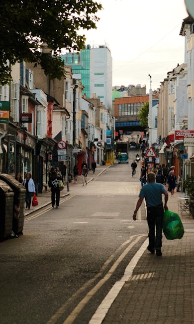 A busy commercial street viewed from a slightly elevated perspective, with a variety of small retail shops and cafes lining both sides of the pavement. The buildings are predominantly two to three sto