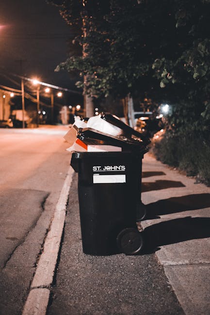 A black wheelie bin labeled 'St. John's' positioned on a sidewalk curbside at night, with the lid open and filled with various waste items including cardboard boxes and plastic packaging. The bin is p
